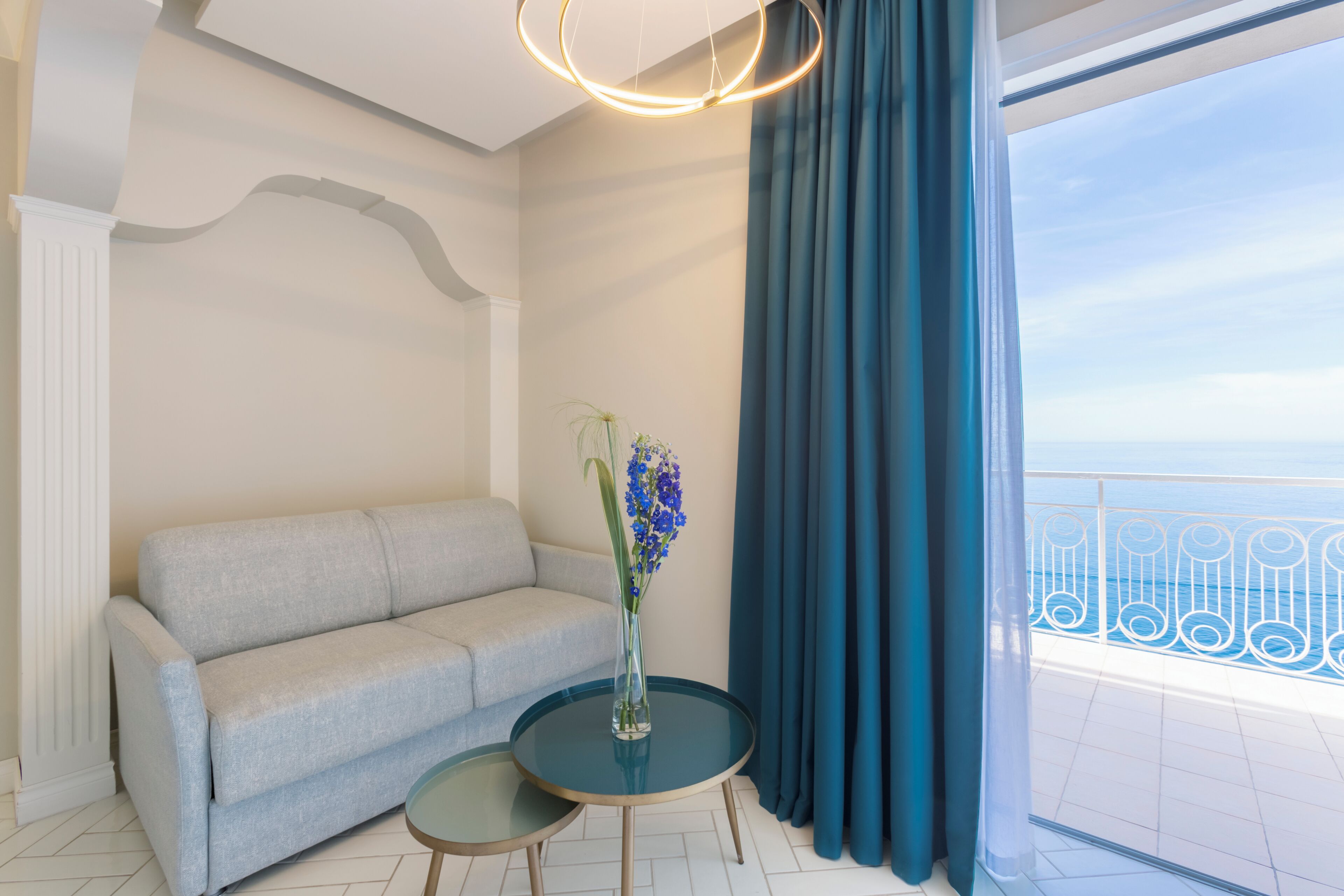A room with a sofa, a small table, a vase with flowers, and a view of the sea through a balcony.
