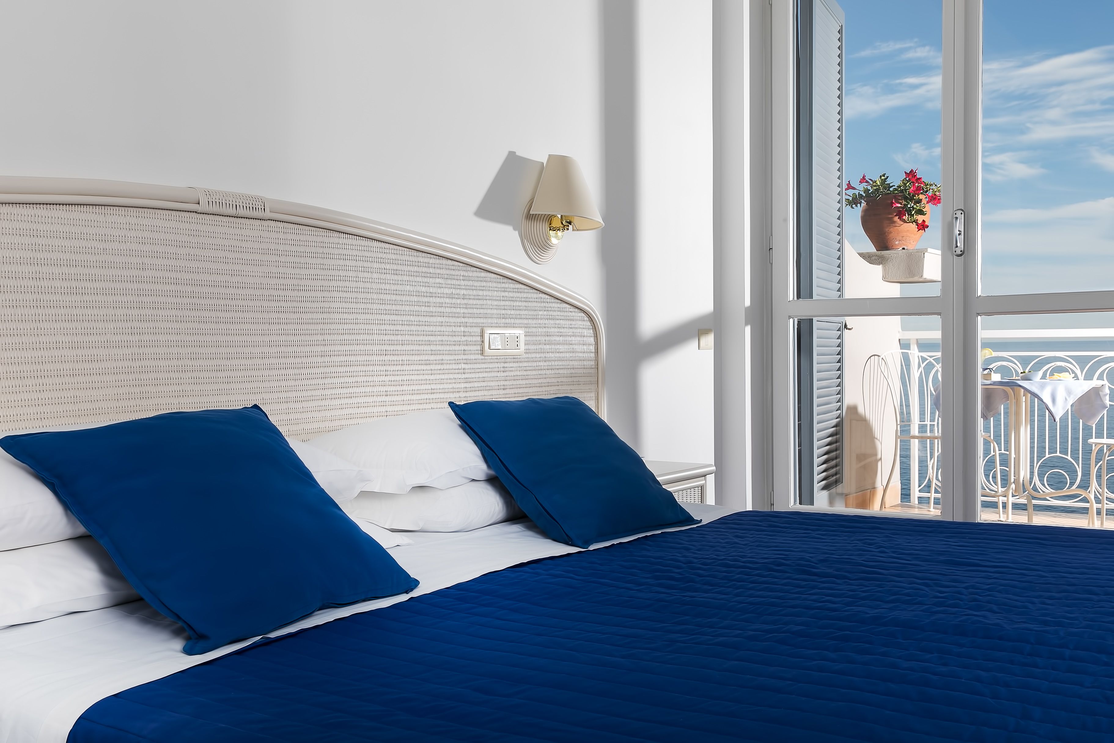 Una cama con ropa de cama azul, cabecera blanca y ventana con vista a un balcón y una planta en maceta.