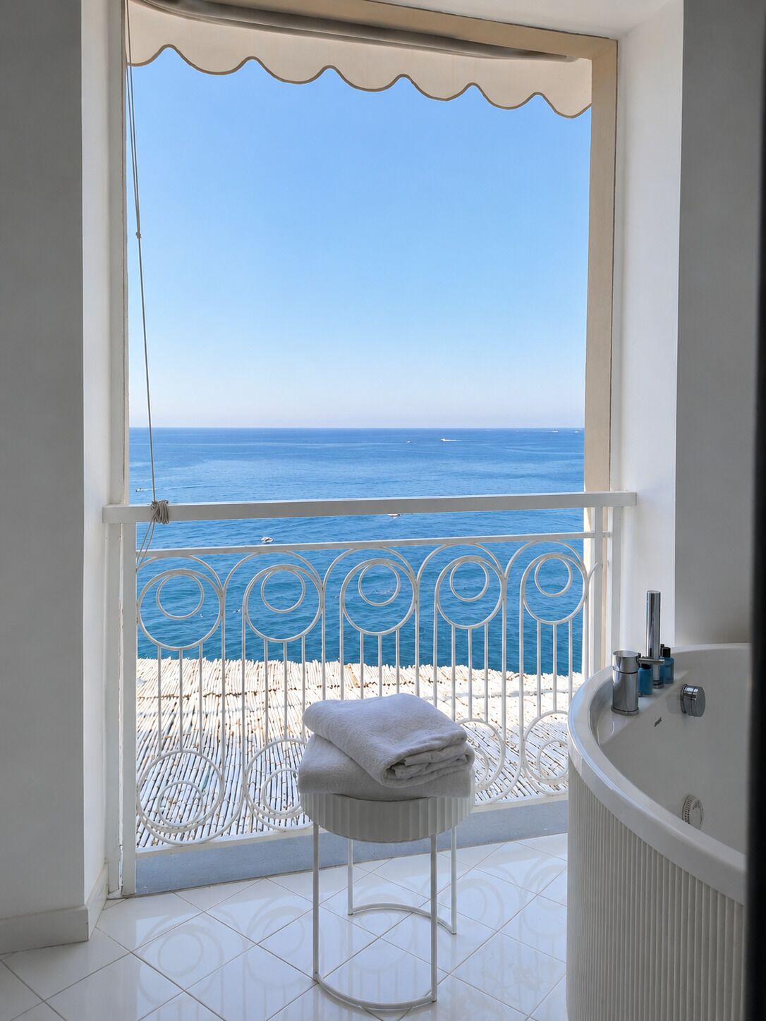 Un balcón con vista al mar, una bañera blanca y una toalla sobre un taburete.