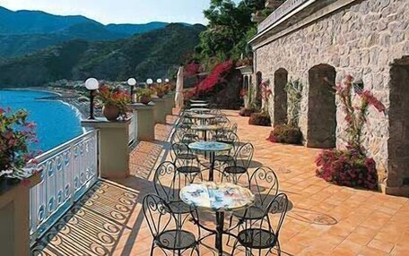 An outdoor terrace with round tables and chairs, overlooking a body of water and hills.