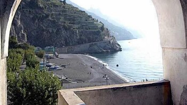 A coastal view through an arched window, featuring a beach, rocky cliffs, and a clear sky. - Palazzo Vingius (Minori)