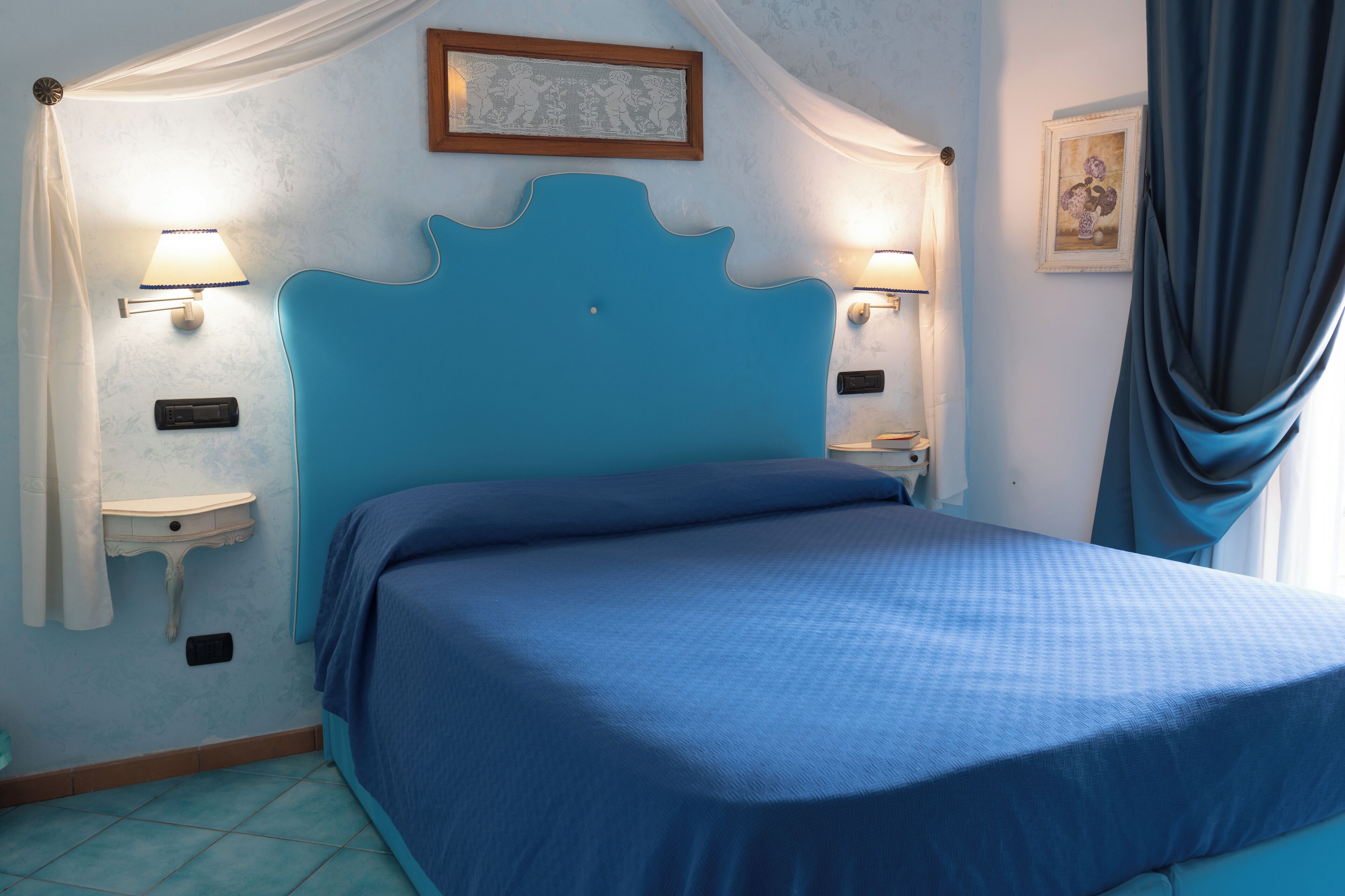 A bedroom with a blue headboard, two bedside tables with lamps, a small bedside table with a radio, and a window with a curtain.