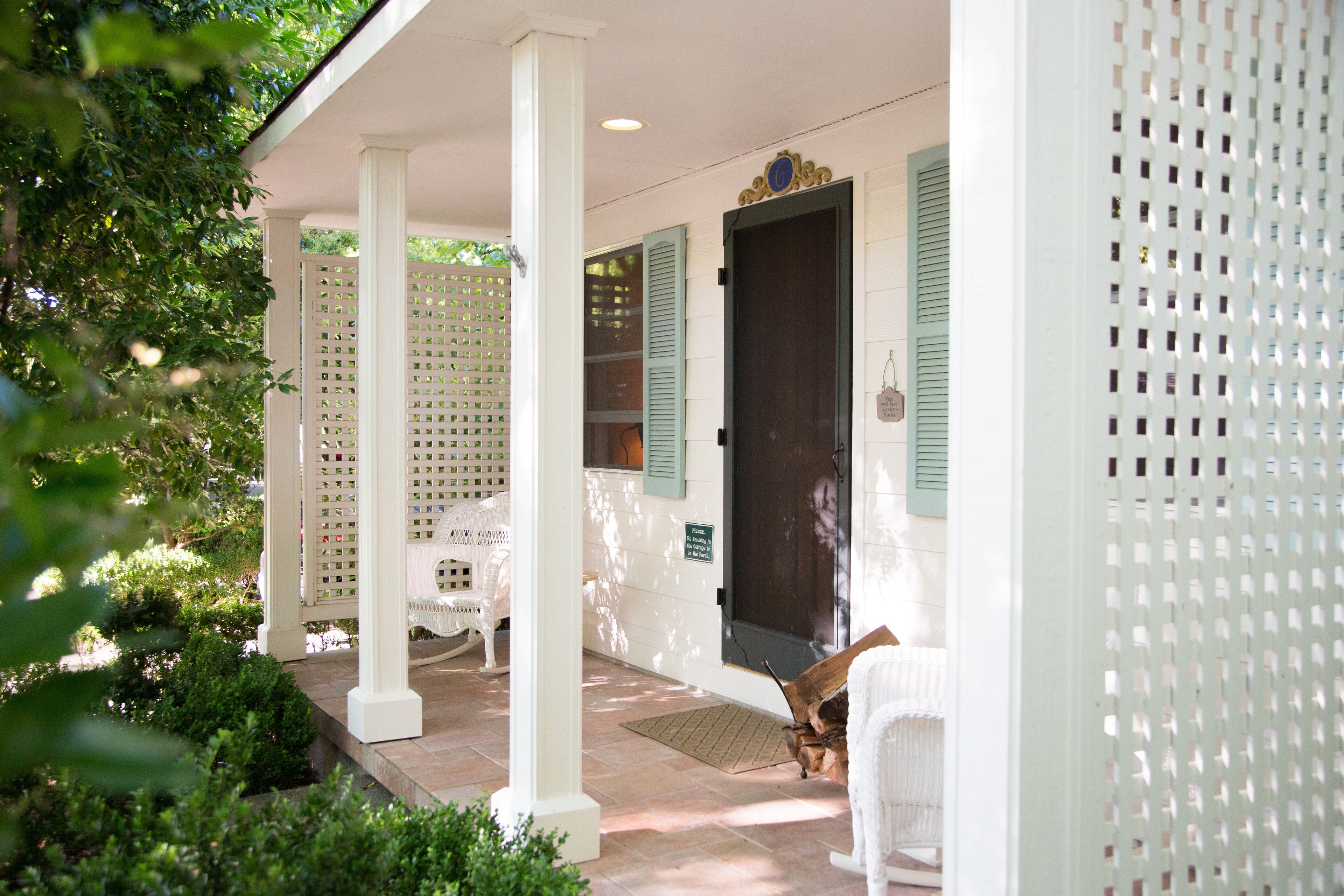 A covered porch with white columns and green shutters, surrounded by greenery.