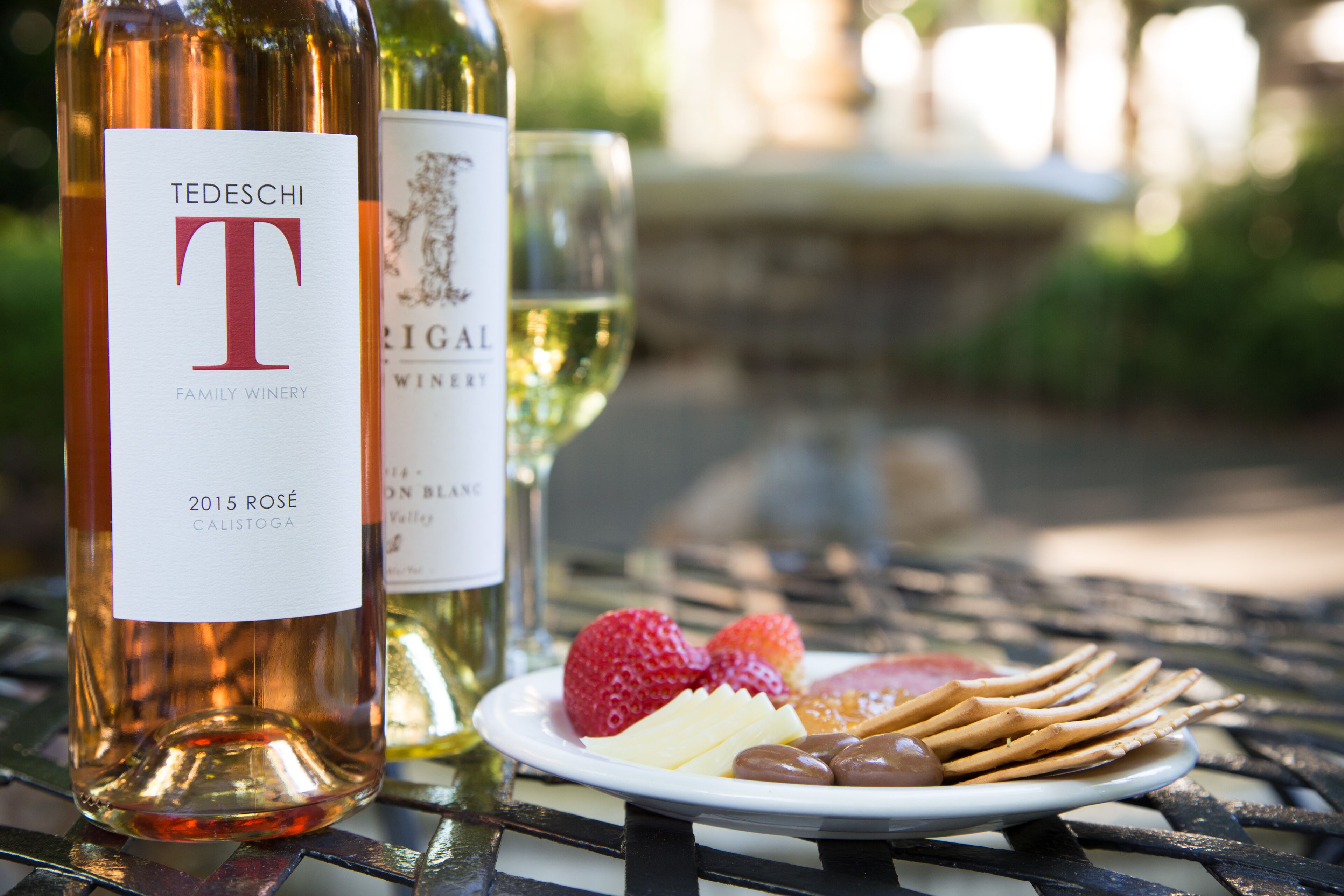 Two wine bottles, a plate with strawberries, cheese, and crackers, and a glass of white wine on a metal table.