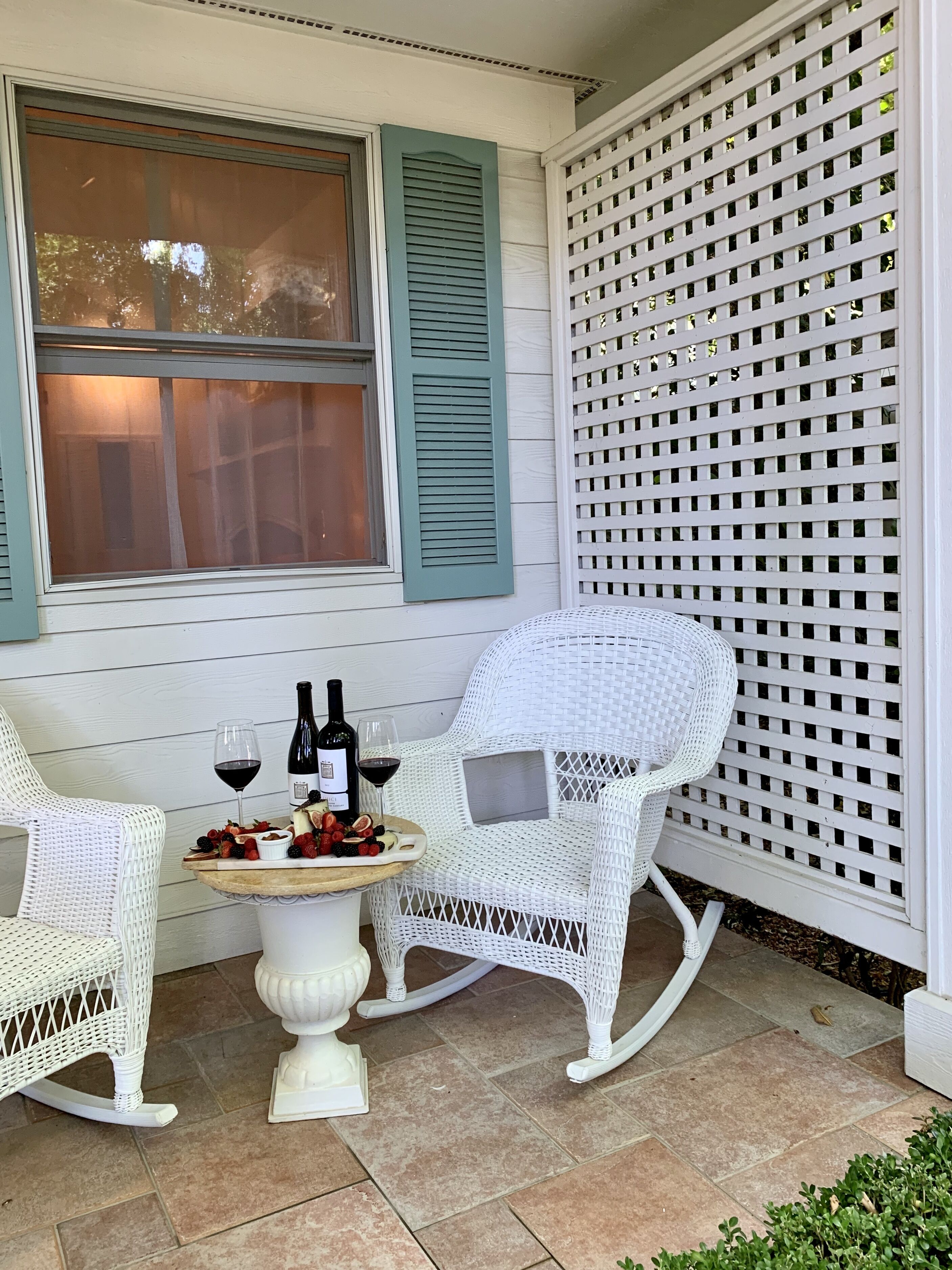 A patio with two white wicker chairs, a small round table, and a bottle of wine.