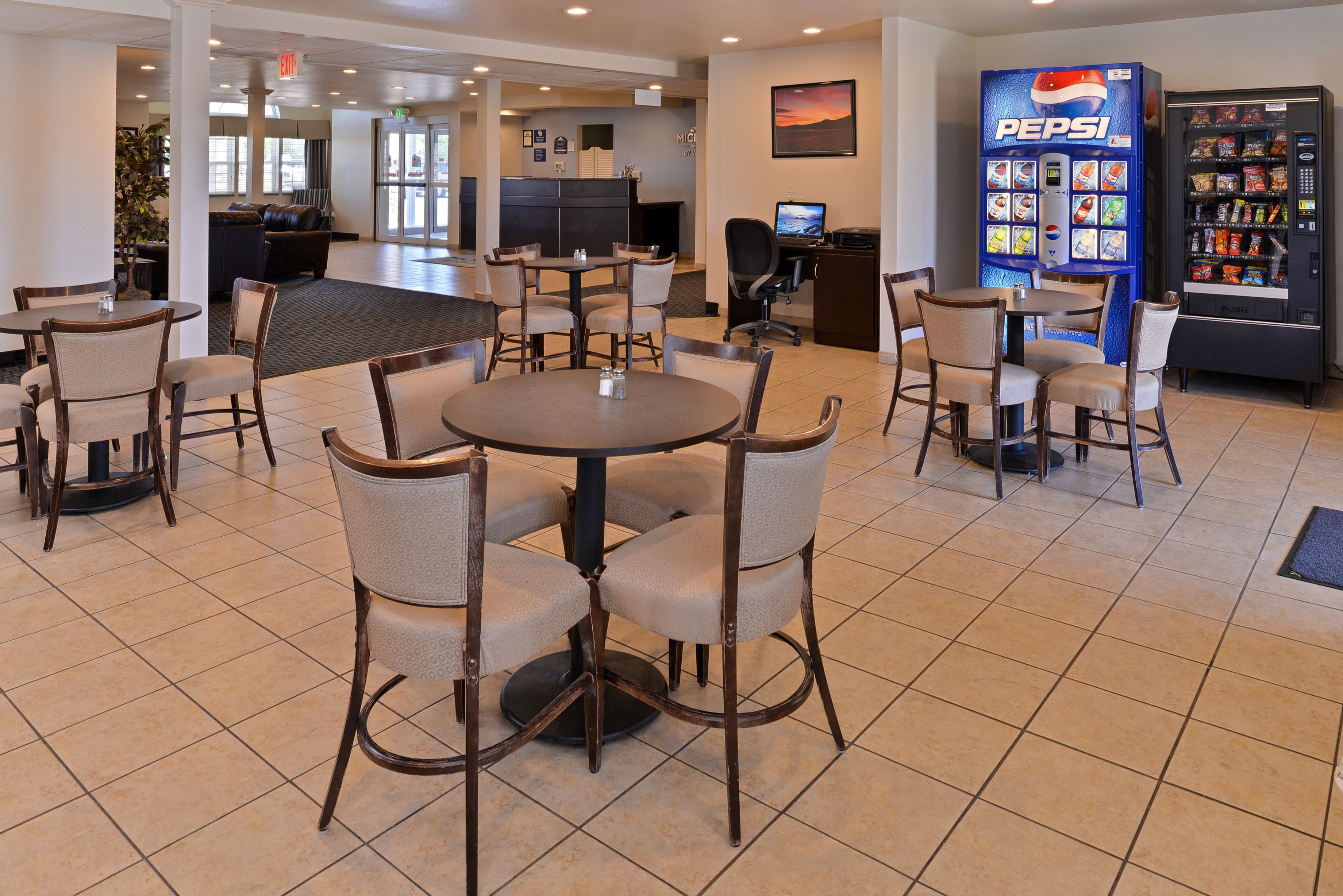 A hotel lobby with round tables and chairs, a reception desk, a computer, and a Pepsi vending machine.