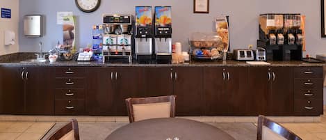 Et morgenmadsområde på et hotel med kaffemaskine, juiceautomater og et ur på væggen.
