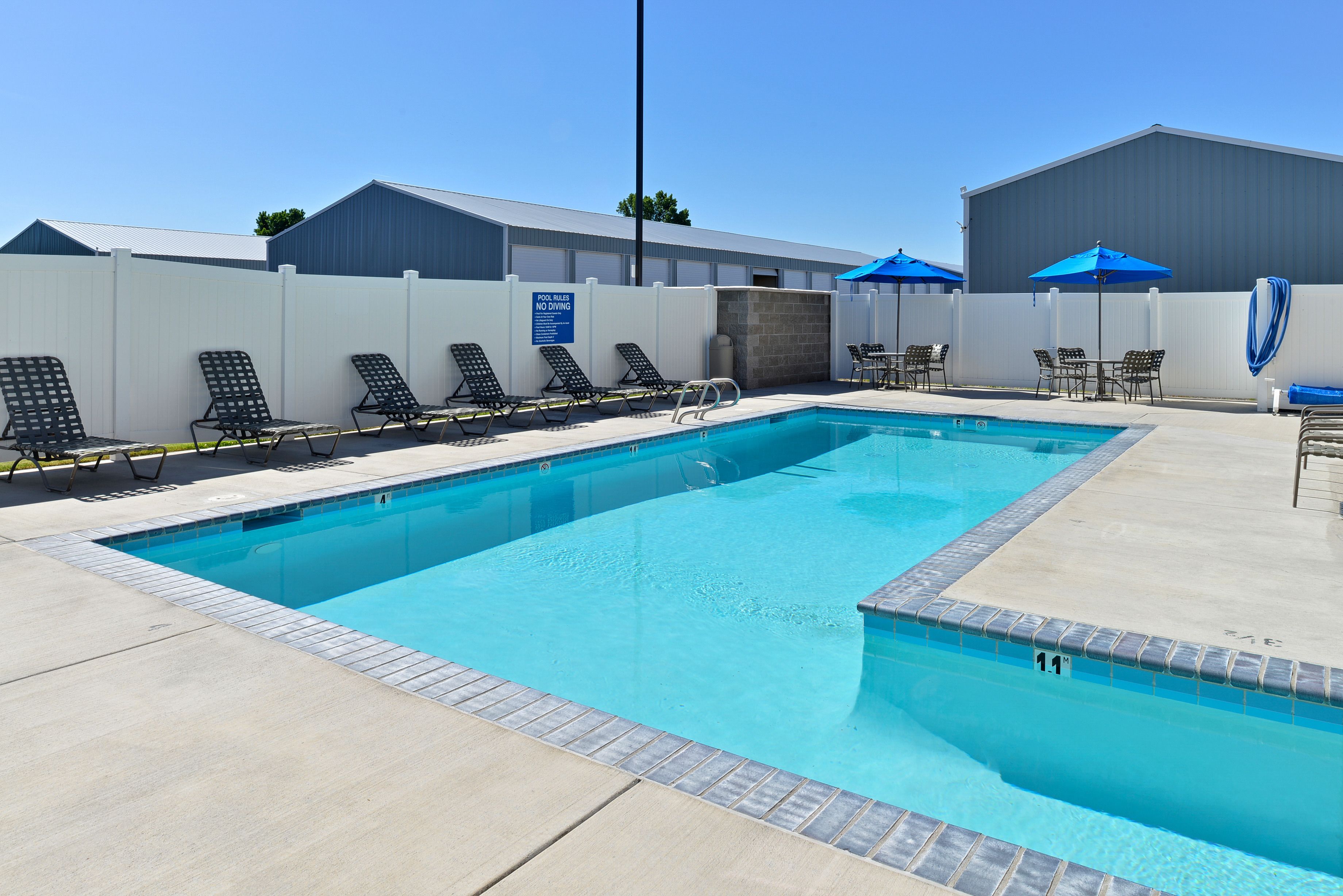 A swimming pool with lounge chairs and umbrellas, surrounded by a white fence and a building.