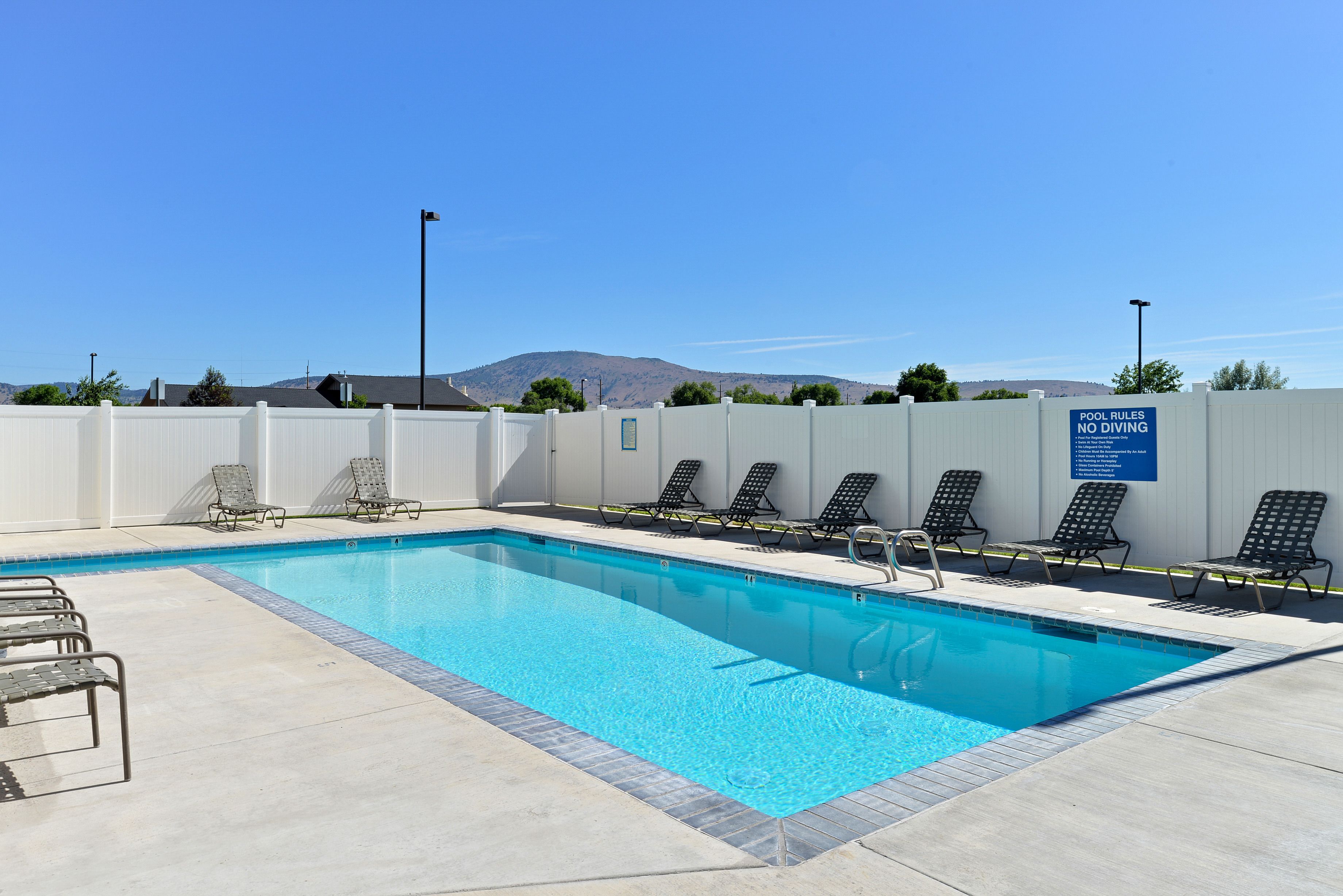 A rectangular swimming pool with a clear blue water surface, surrounded by a white fence and several black lounge chairs. A sign on the fence states "POOL RULES NO DIVING."