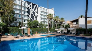 Una piscina con sillas de descanso y sombrillas, rodeada de palmeras y un edificio moderno con balcones.