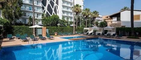 A swimming pool with lounge chairs and umbrellas, surrounded by palm trees and a modern building with balconies.