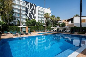 Una piscina con sillas de descanso y sombrillas, rodeada de palmeras y un edificio moderno con balcones.