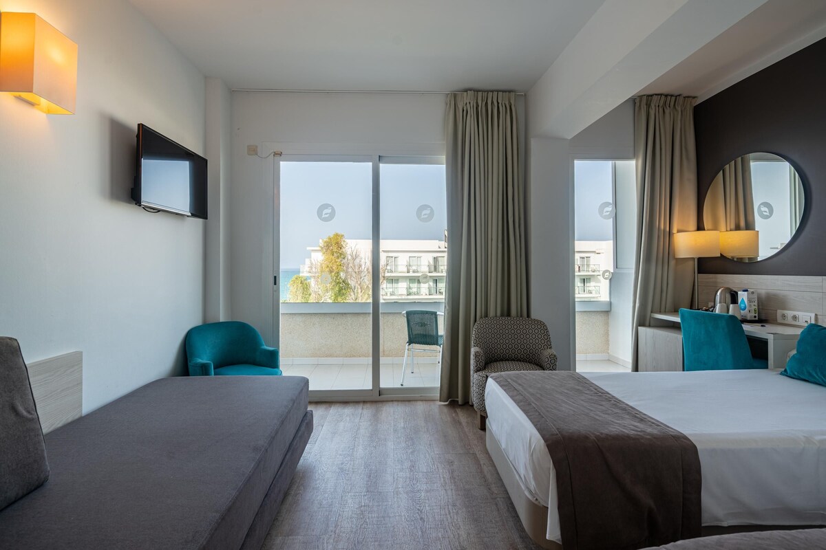 Habitación de hotel con dos camas, televisión, escritorio y balcón con vistas.