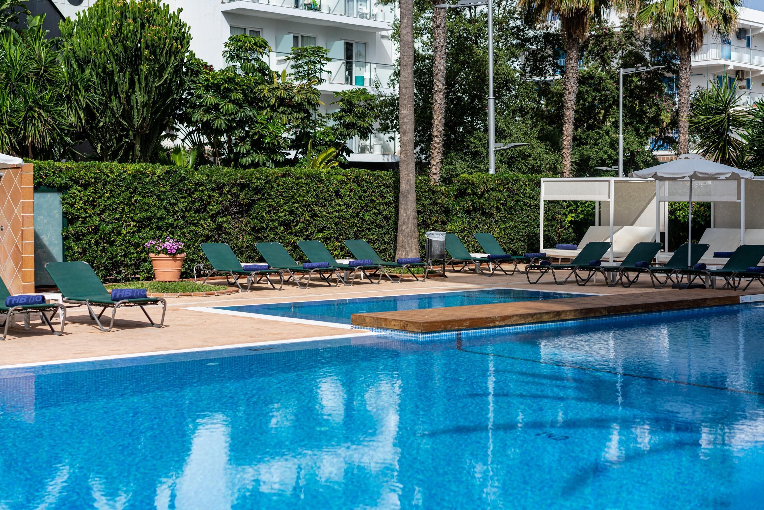 A swimming pool with lounge chairs and a covered seating area, surrounded by greenery and buildings.
