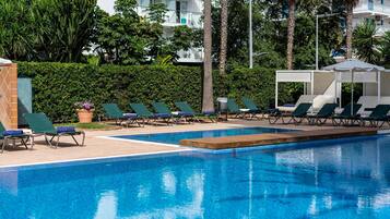 A swimming pool with lounge chairs and a covered seating area, surrounded by greenery and buildings.