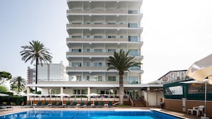 Un hotel de varios pisos con piscina, palmeras y mobiliario de exterior.