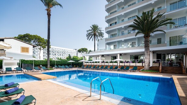 Una piscina con sillas de descanso y sombrillas, rodeada de palmeras y un edificio moderno.
