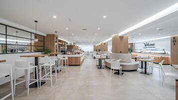 Un café moderno con taburetes blancos, mesas redondas y una zona central con sofás.