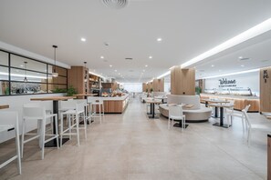 Un café moderne avec des tabourets de bar blancs, des tables rondes et un espace salon central agrémenté d’un canapé.
