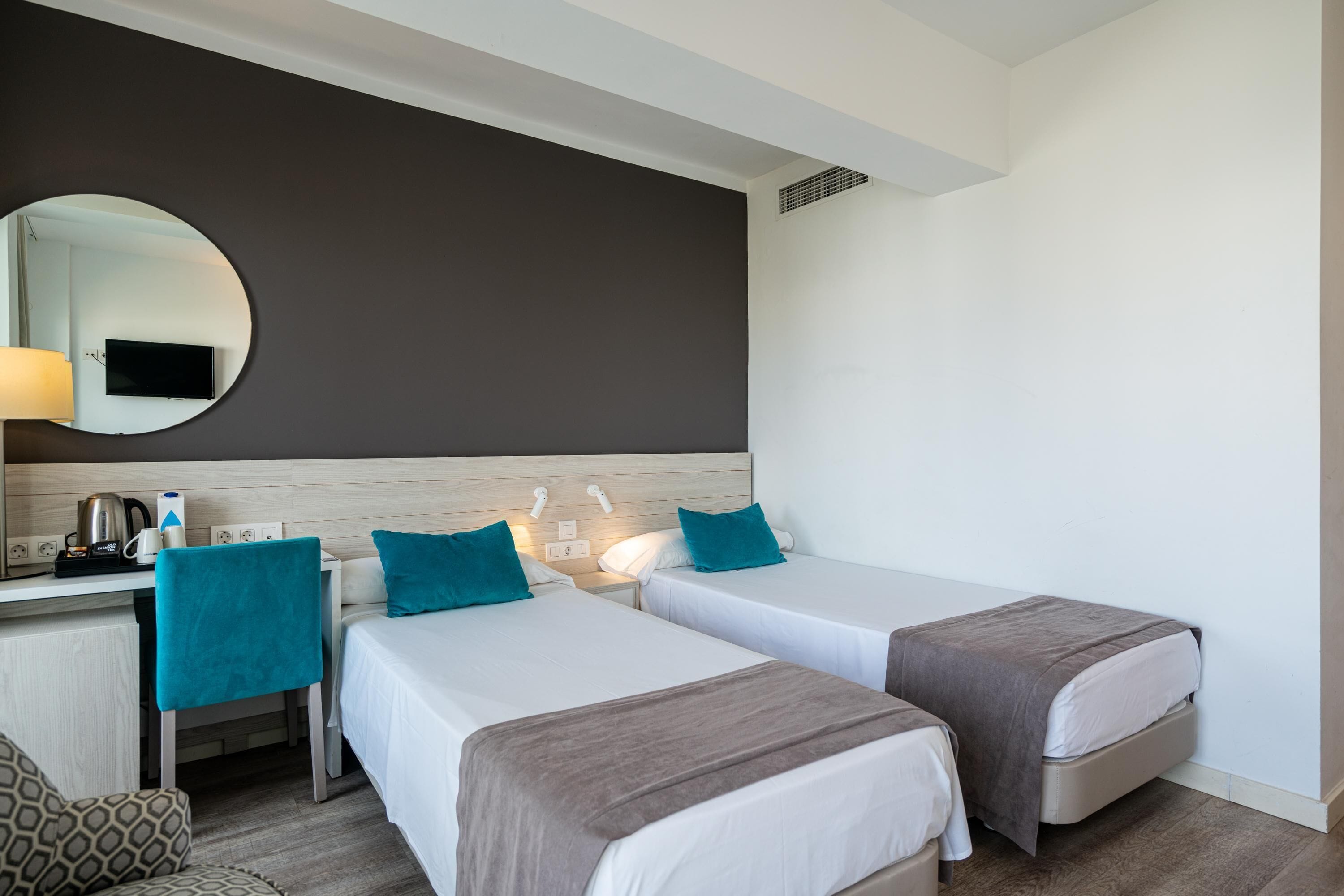 Una habitación de hotel con dos camas individuales, un escritorio con silla, un espejo y un televisor en la pared.