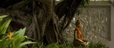 A Buddha statue under a large tree with intricate wall carvings in the background.