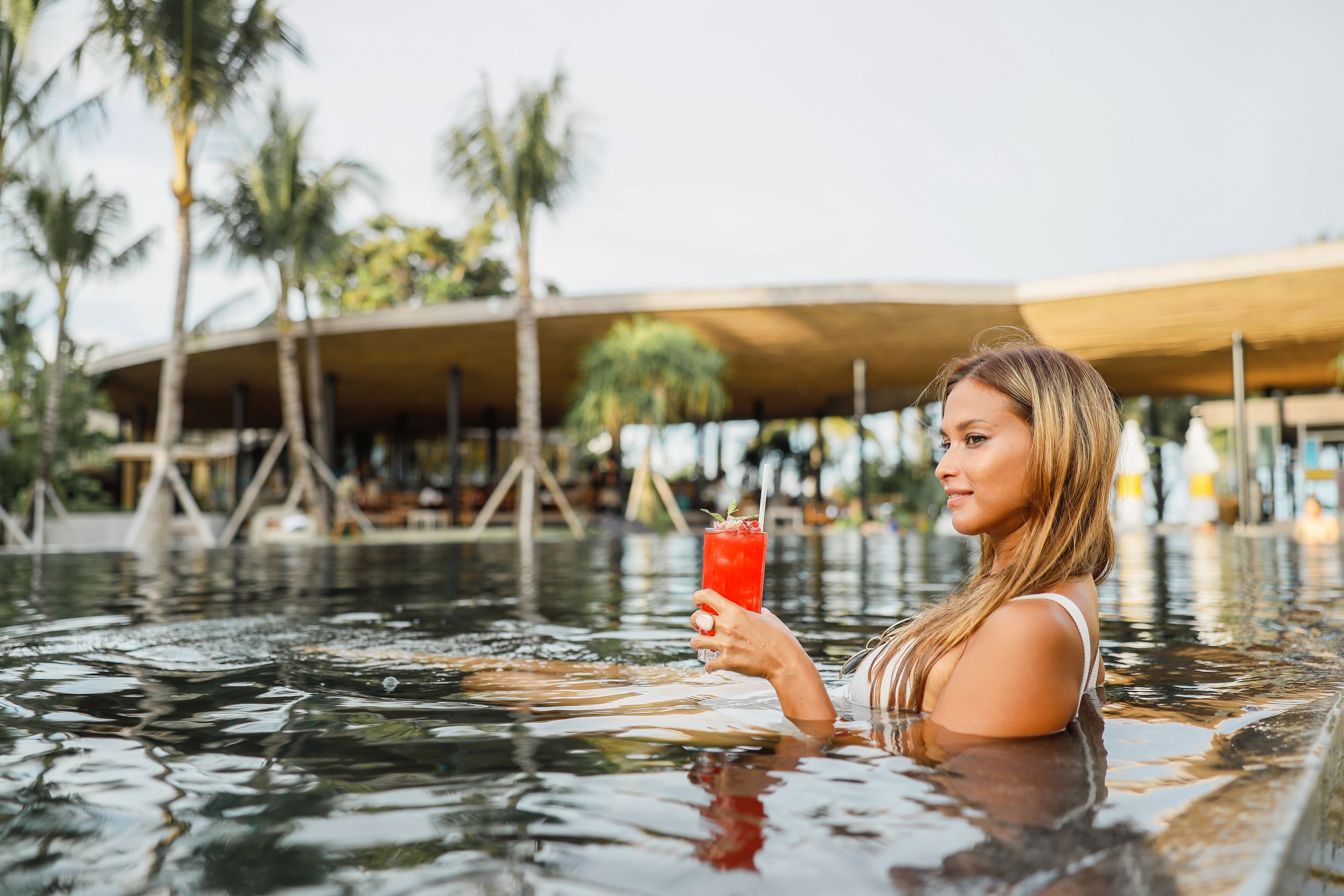 A person in a swimming pool holding a drink, with palm trees and a covered structure in the background.