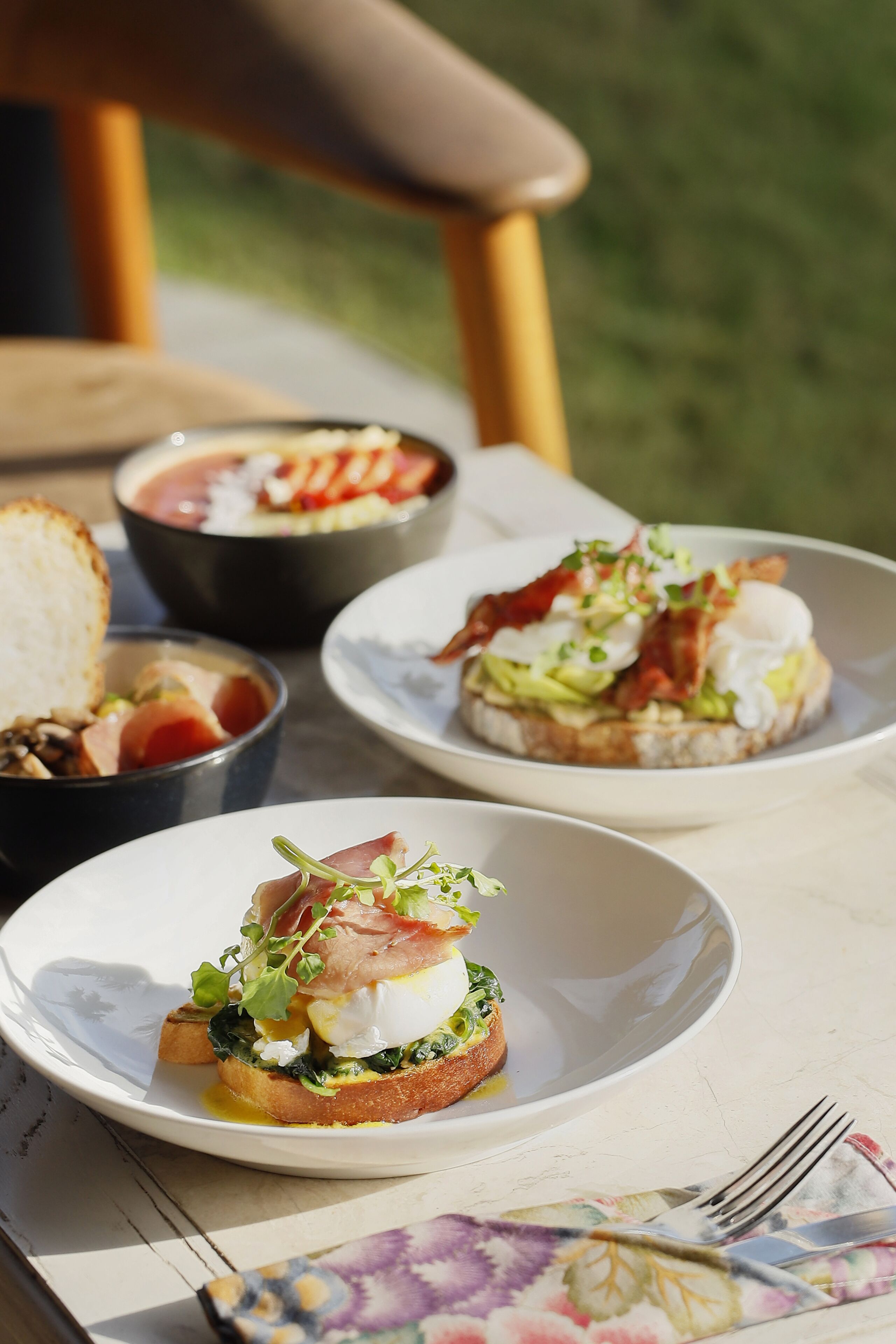 A table with three plates of food, including toast with greens and eggs, and bowls of soup and salad.