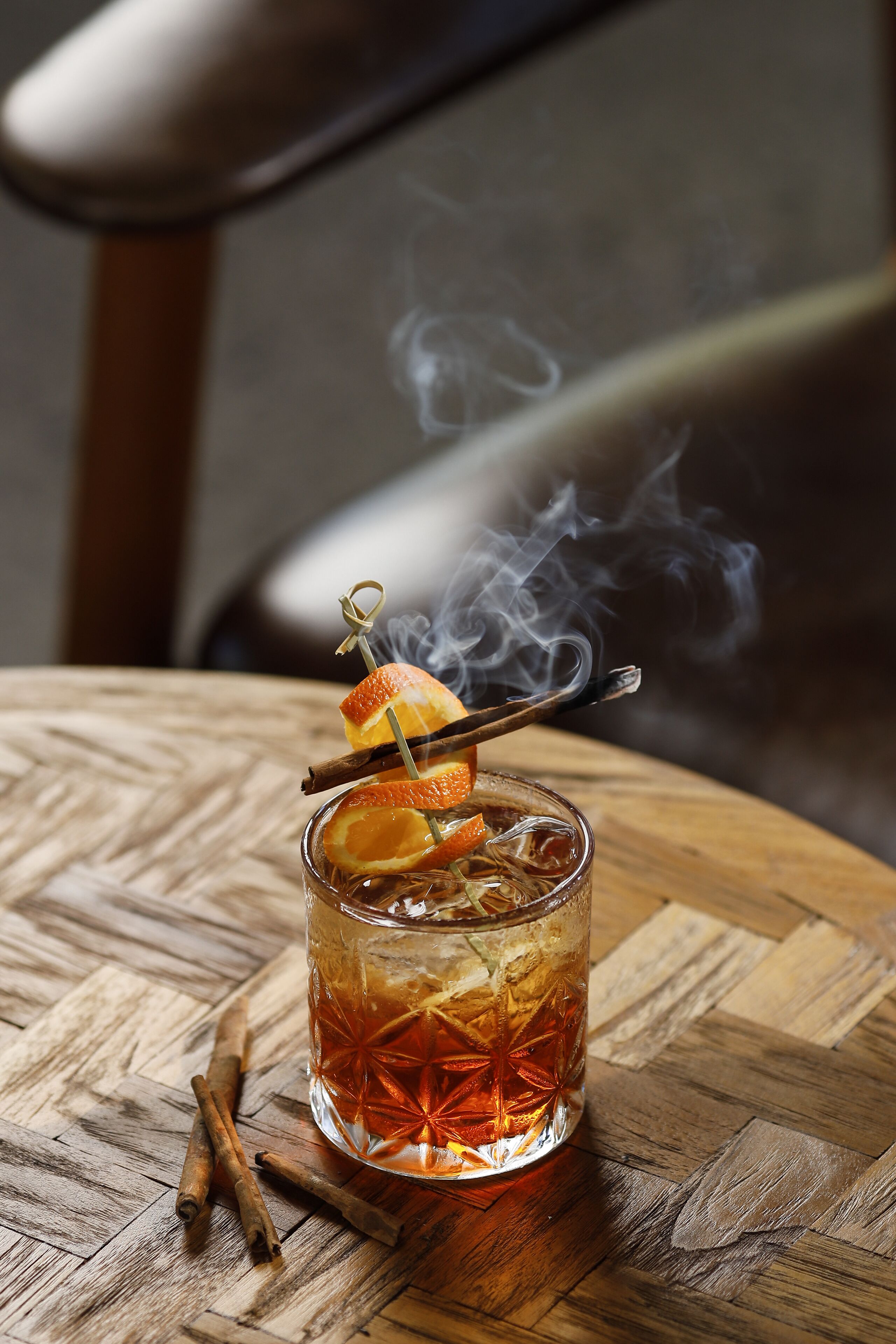 Un vaso de bebida color ámbar adornado con una rama de canela y una rodaja de naranja, con vapor ascendiendo desde la bebida, sobre una mesa de madera con textura tejida.