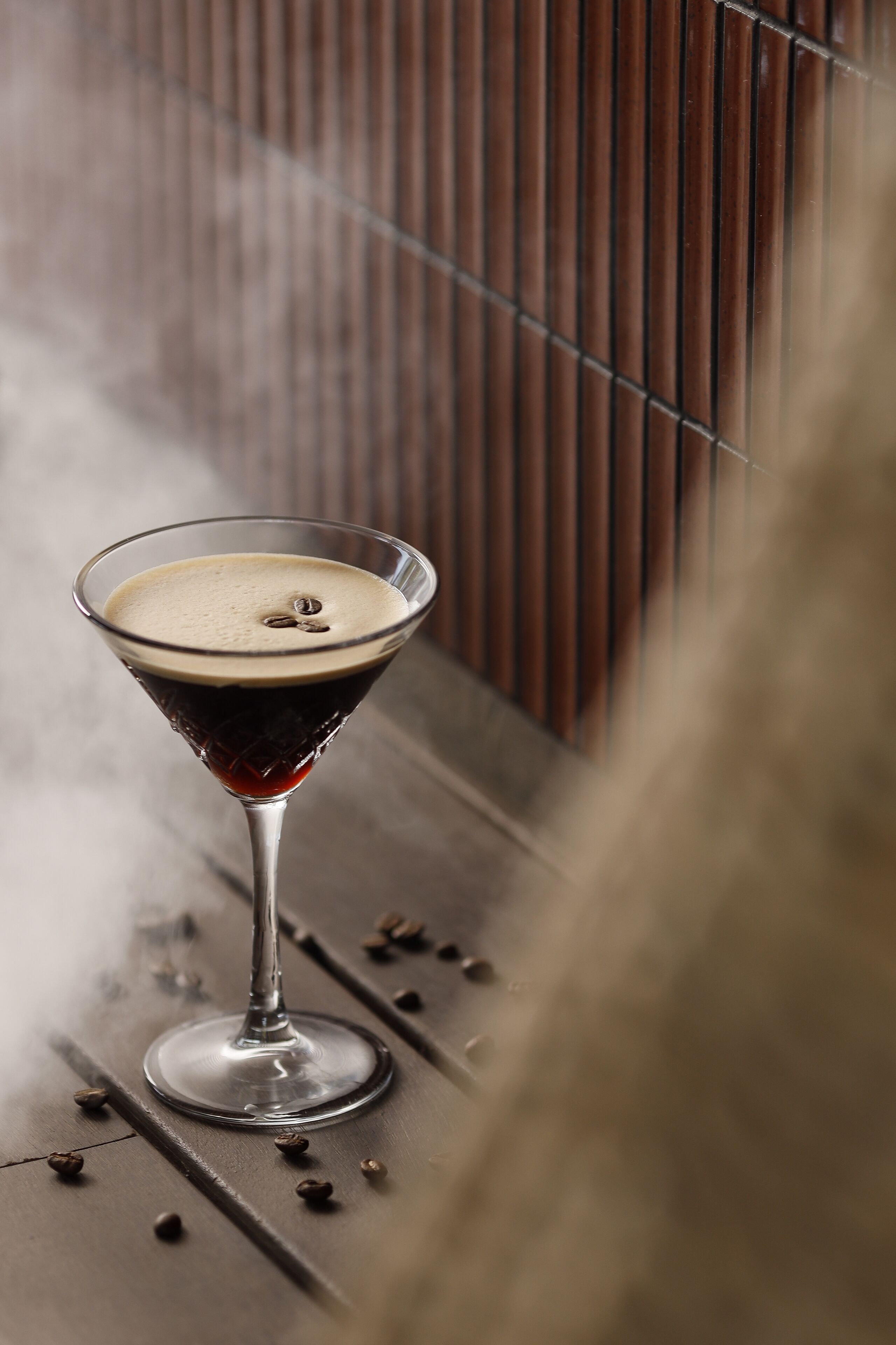 A martini glass with a dark liquid and a frothy top, placed on a wooden surface with scattered coffee beans.