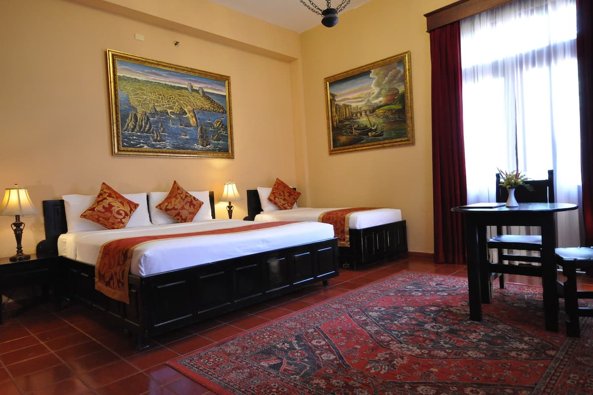 Een hotelkamer met twee bedden, een bureau en een stoel. Er hangen twee schilderijen aan de muur, een rood gordijn en een kamerbreed tapijt.
