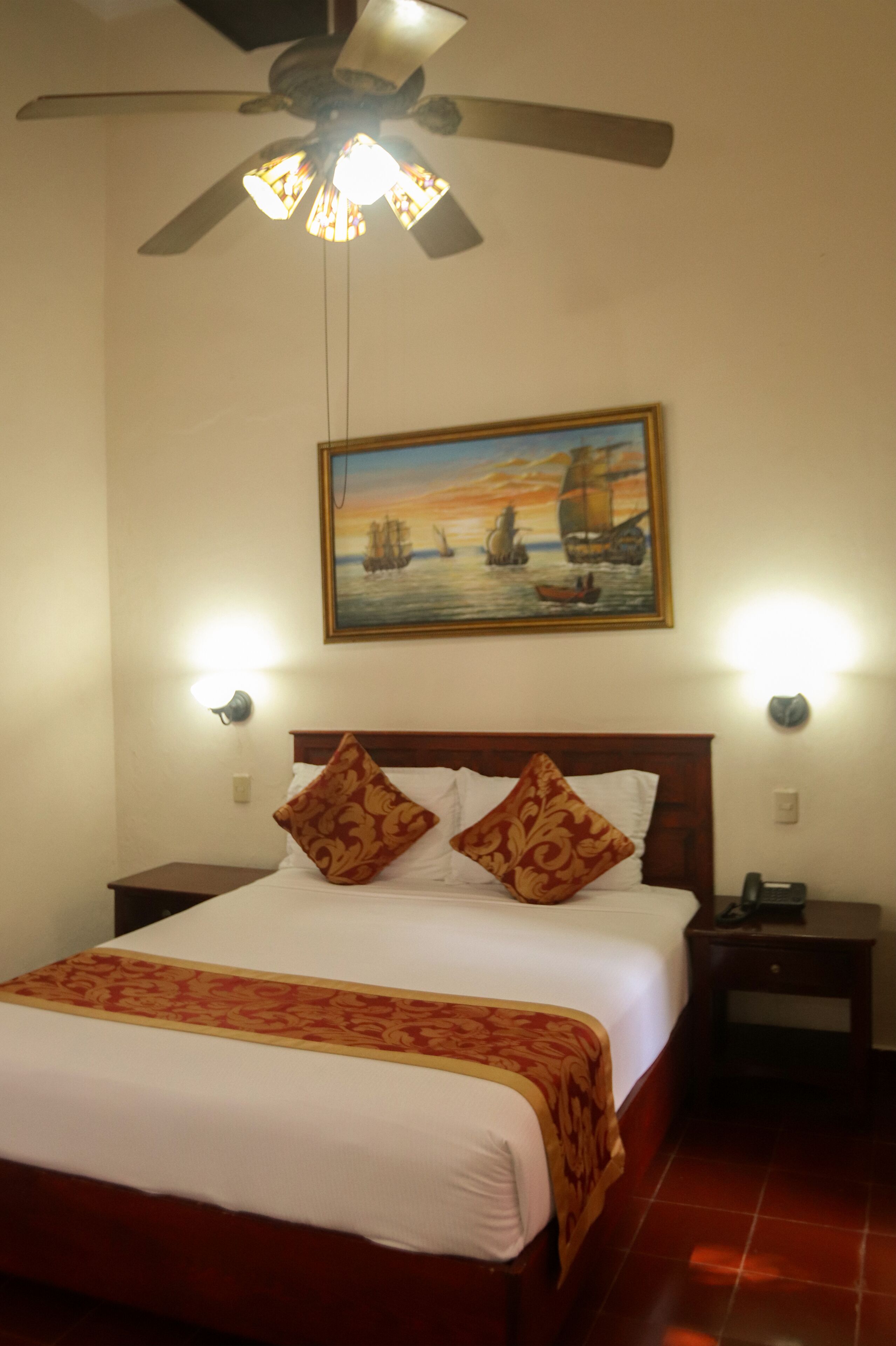 A hotel room with a bed, a ceiling fan, a painting of ships on the wall, and two wall-mounted lights.