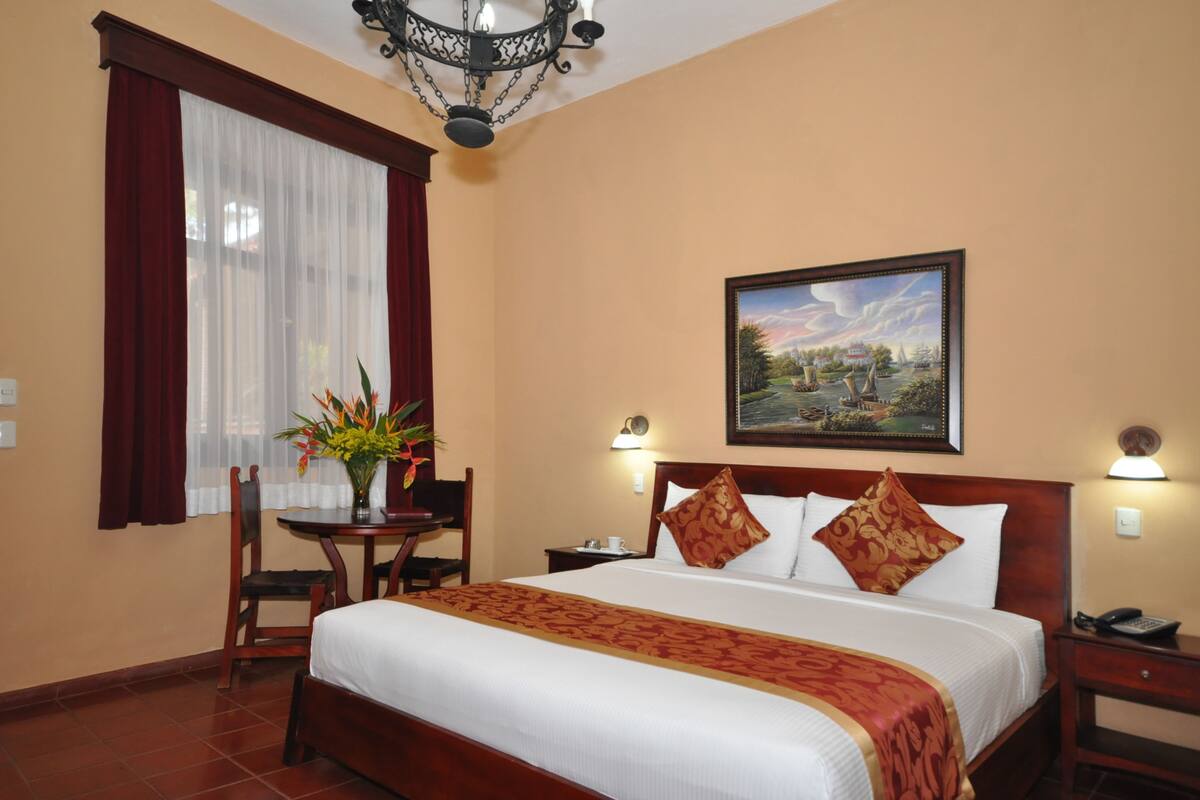 Een hotelkamer met een groot bed, een bureau met stoel, een bloevas, een schilderij aan de muur en een kroonluchter.