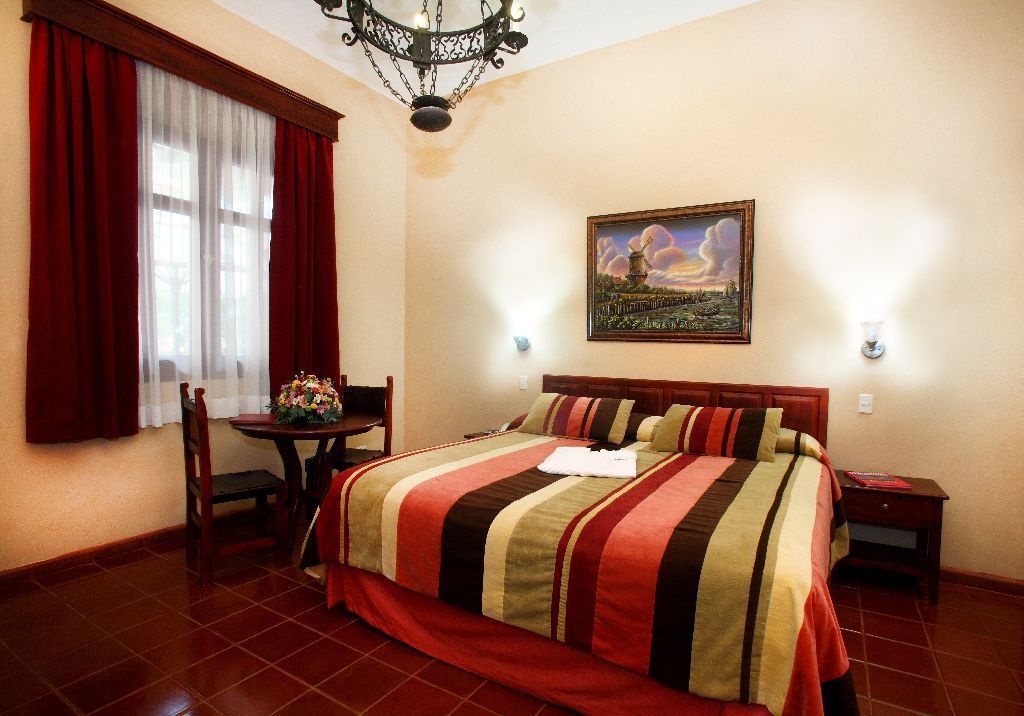 A hotel room with a large bed, a wooden nightstand, a desk with a chair, and a painting on the wall.