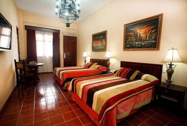 Habitación de hotel con tres camas, una mesa de comedor, sillas, una araña y dos cuadros en la pared.