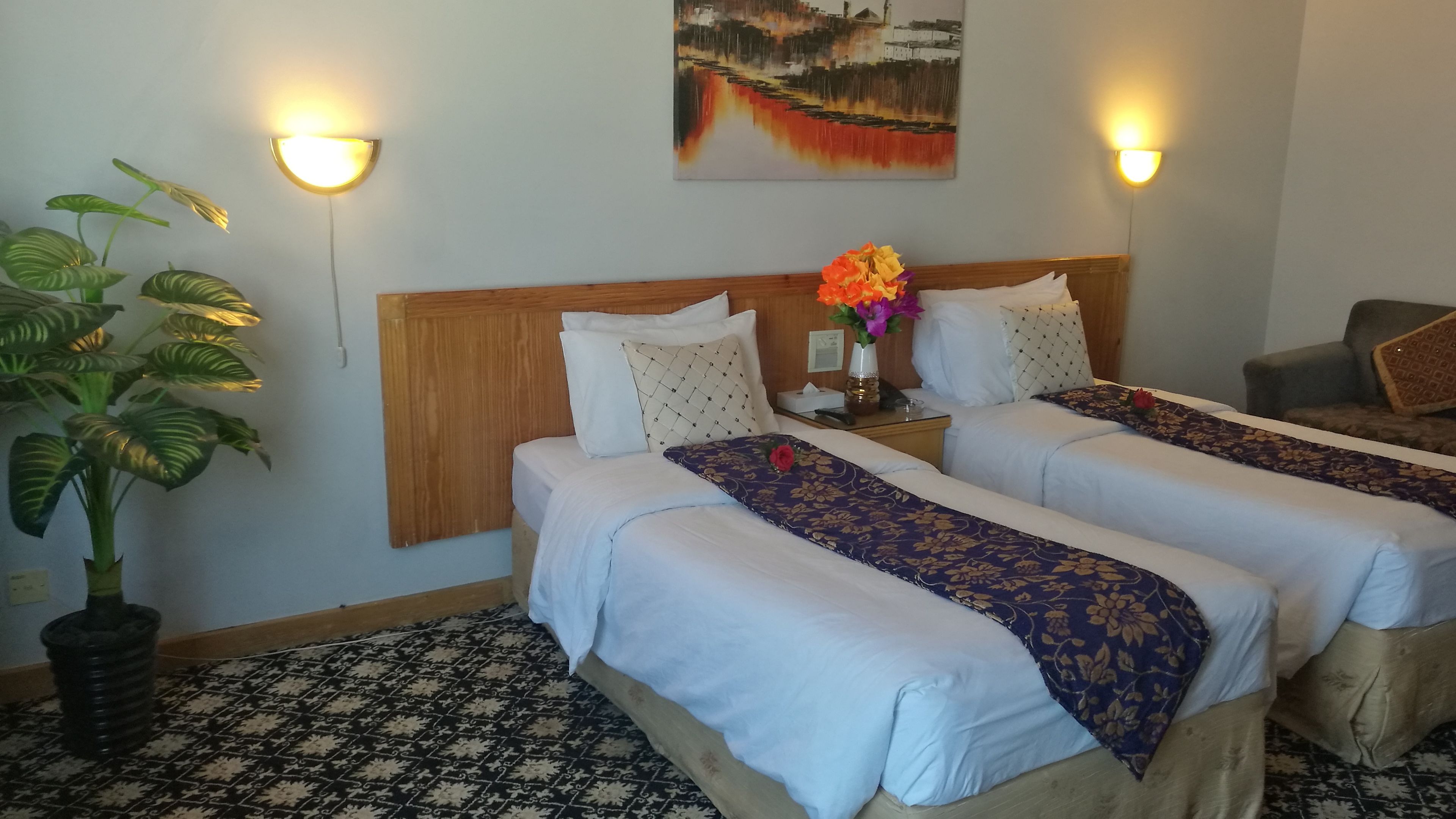 Habitación de hotel con dos camas, una planta grande en maceta, un cuadro en la pared y dos lámparas de pared.