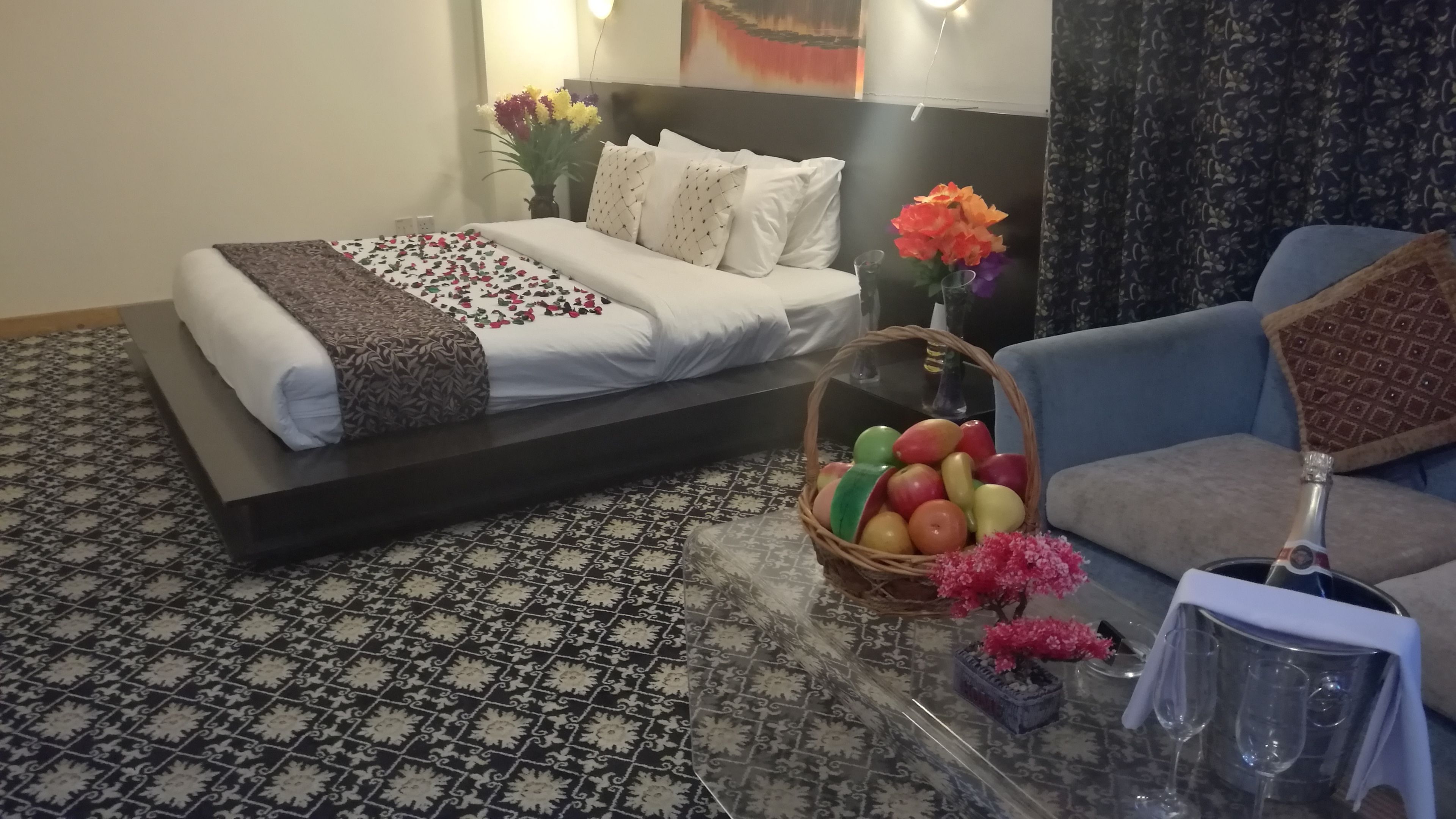 A hotel room with a bed, a sofa, a table with a fruit basket and a vase of flowers, and a bottle of champagne.
