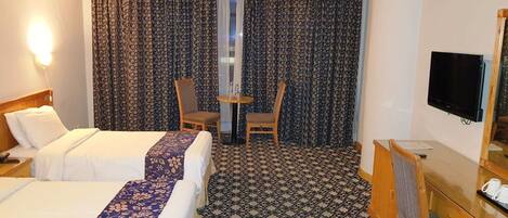 A hotel room with two beds, a desk, a chair, a TV, and patterned curtains.