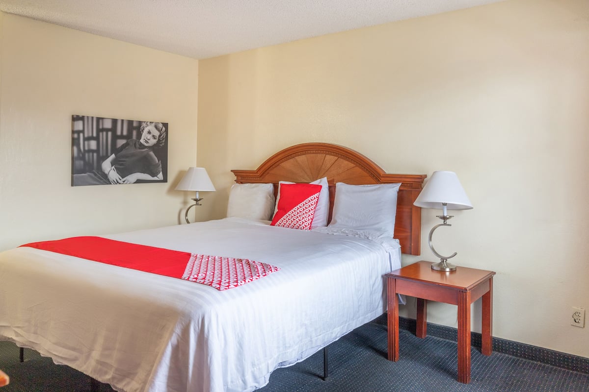 Quarto de hotel com uma cama, duas mesas de cabeceira, uma colcha vermelha e branca, e um quadro na parede.