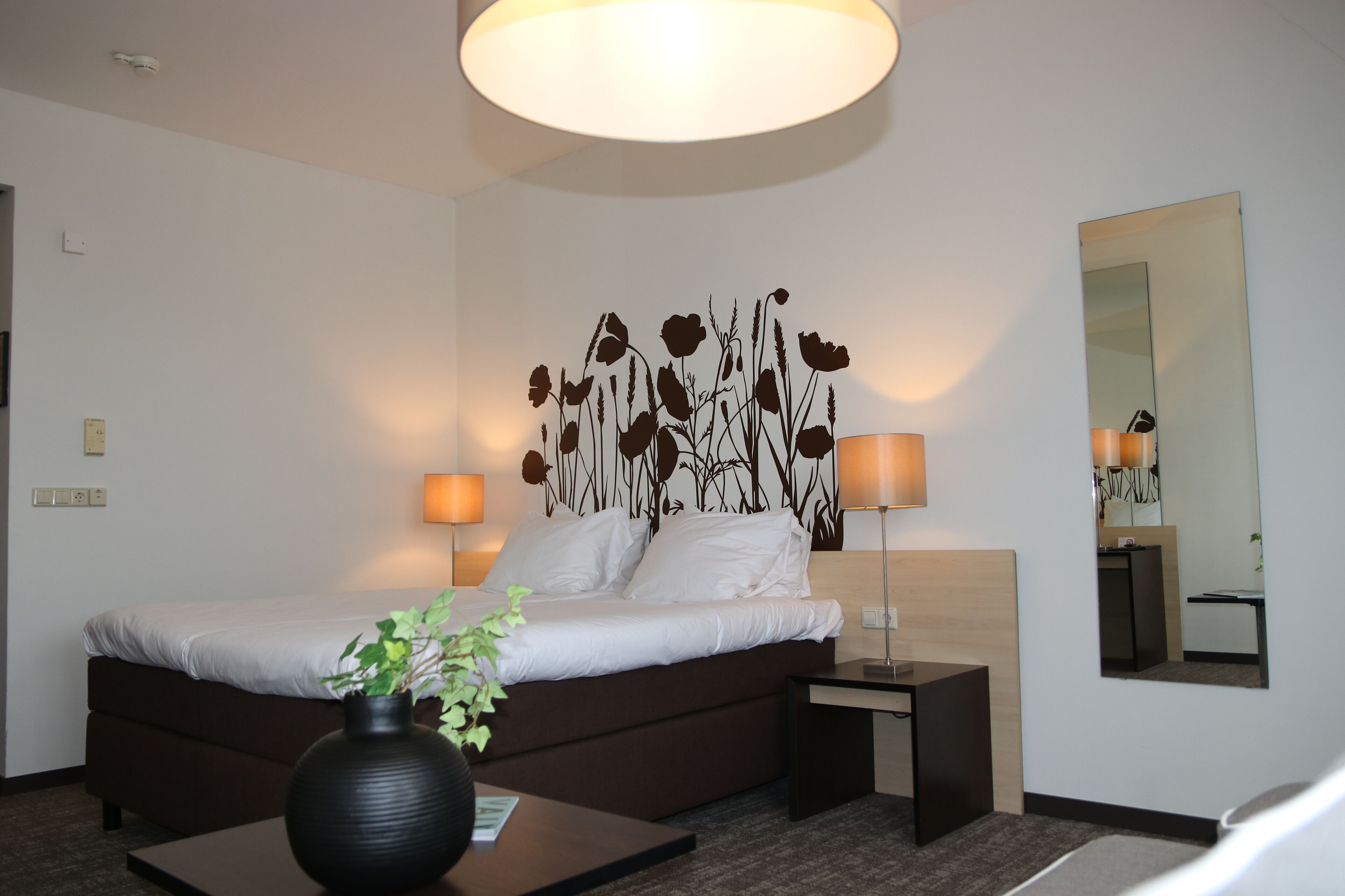 A modern hotel room with a large bed, bedside lamps, a vase with a plant, and a mirror on the wall.