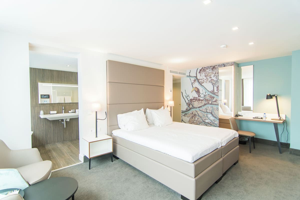 A modern hotel room with a large bed, a desk, a chair, and a bathroom visible through an open door.
