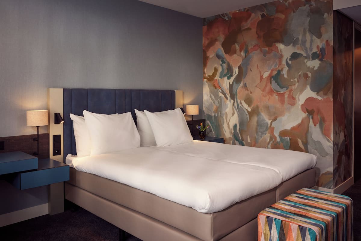 A hotel room with a large bed, two bedside tables with lamps, a colorful abstract wall, and a striped ottoman.