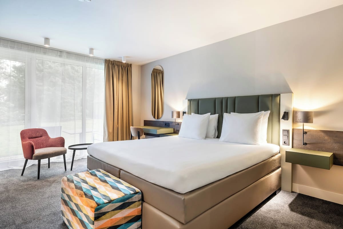 A hotel room with a large bed, a small table, a chair, a bench, and a nightstand with a lamp.