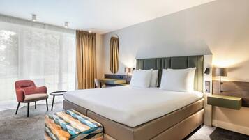 A hotel room with a large bed, a small table, a chair, a bench, and a nightstand with a lamp.