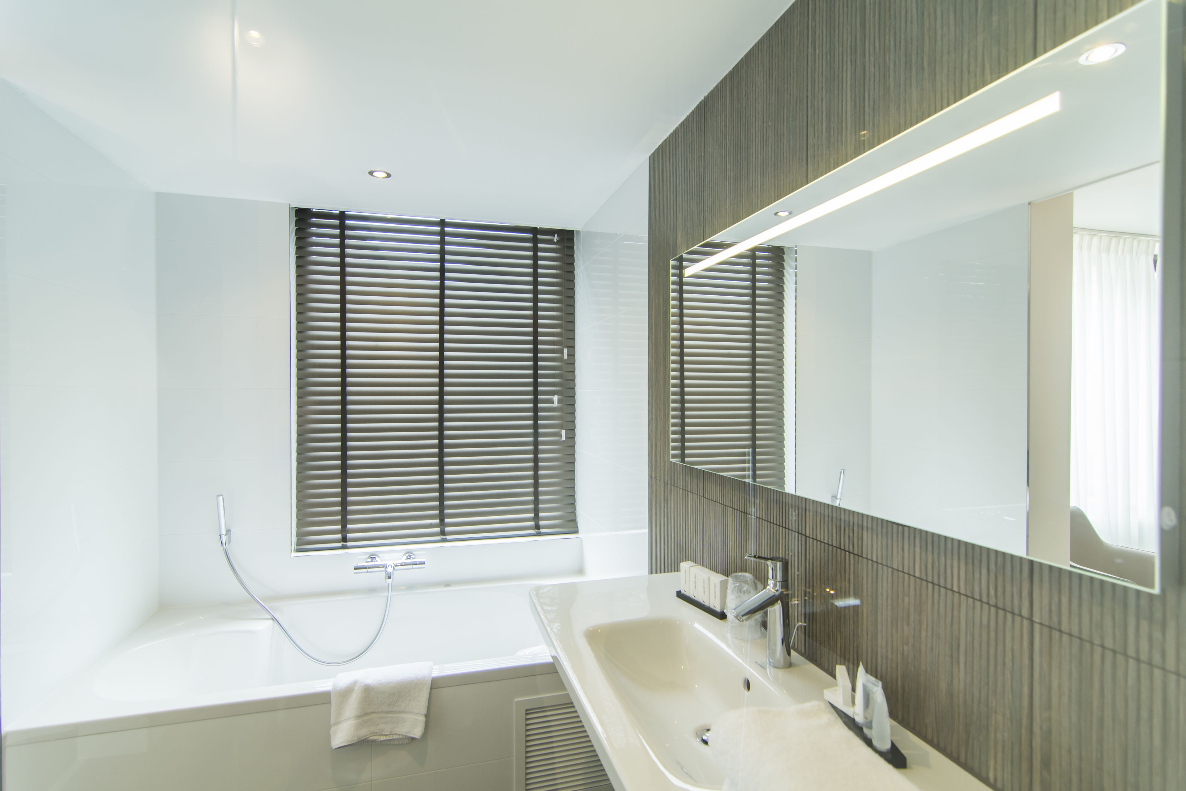 Ein modernes Badezimmer mit Badewanne, großem Spiegel und einem Fenster mit Jalousien.