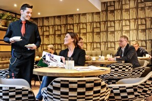 A modern restaurant with checkered chairs, a waiter serving food, and a bookshelf in the background. - Van der Valk Hotel Hilversum - de Witte Bergen (Eemnes)