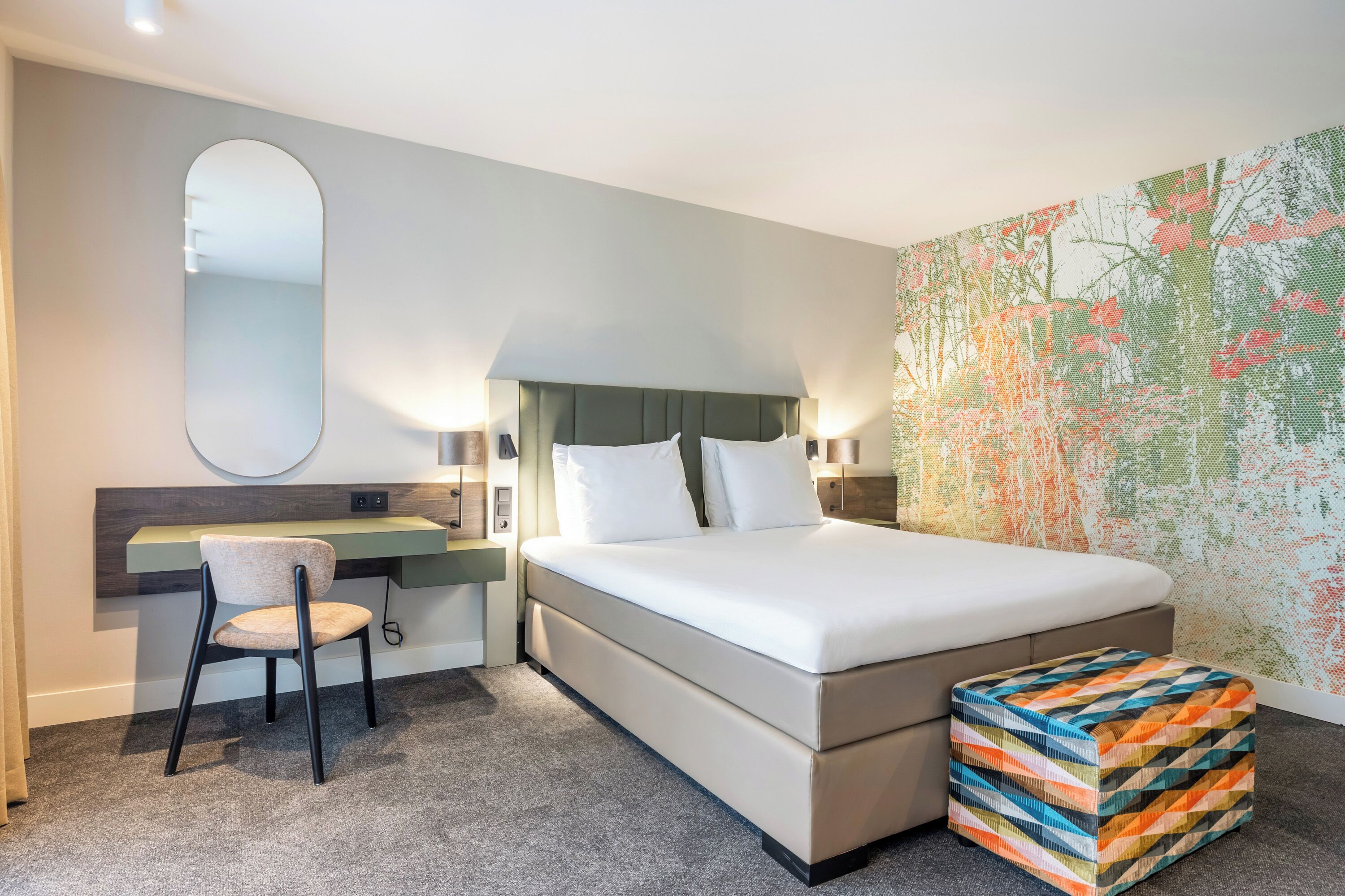 Una habitación de hotel con cama, escritorio, silla, un mural con mapa y un otomano colorido.