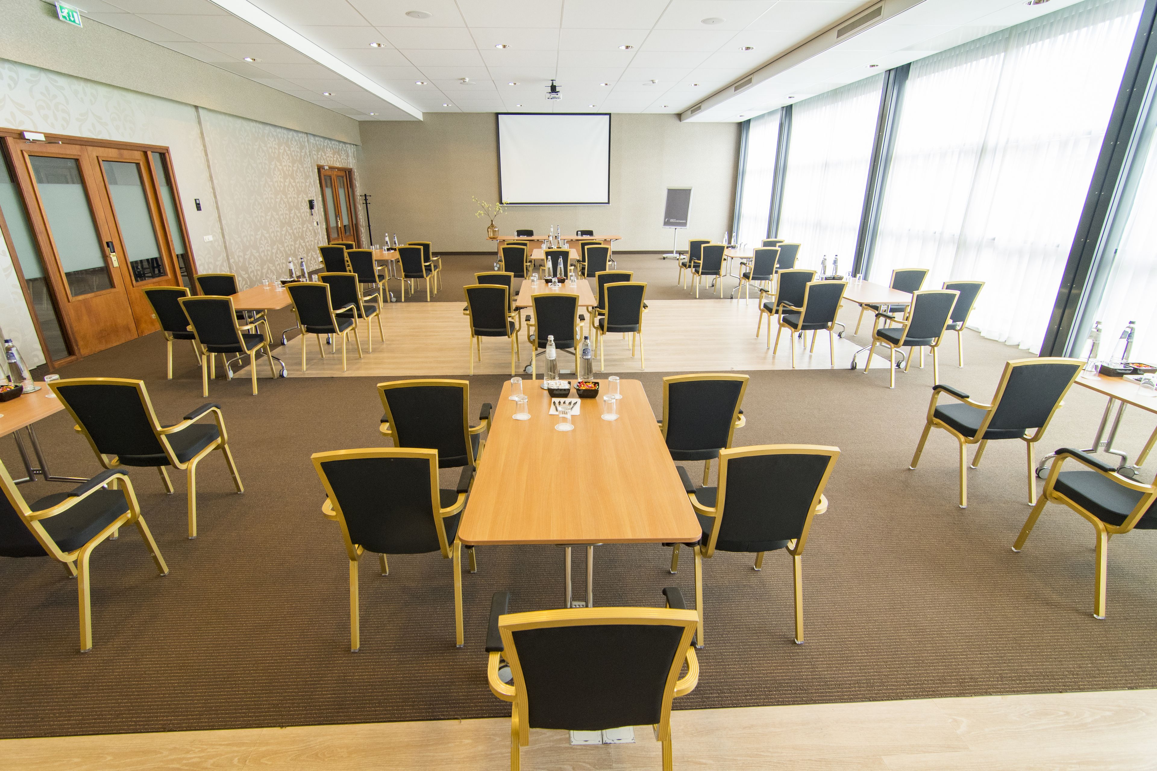 Een vergaderruimte met meerdere tafels en stoelen, een projectiescherm en een groot raam.