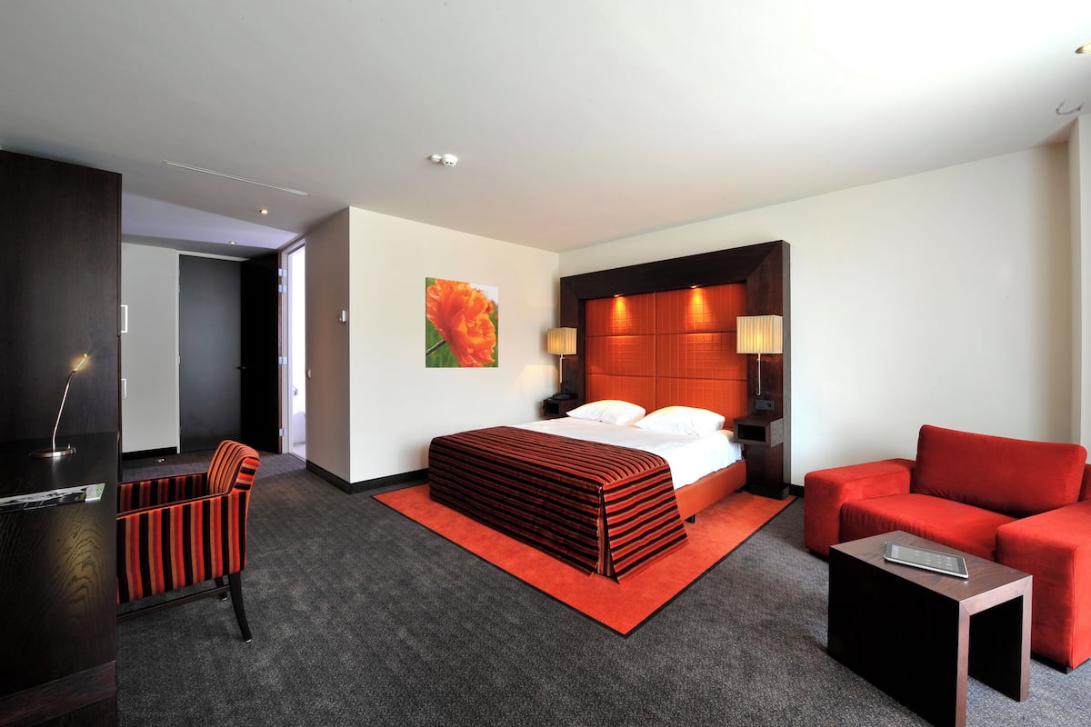 A hotel room with a bed, a red sofa, a small table, a desk, and a chair.