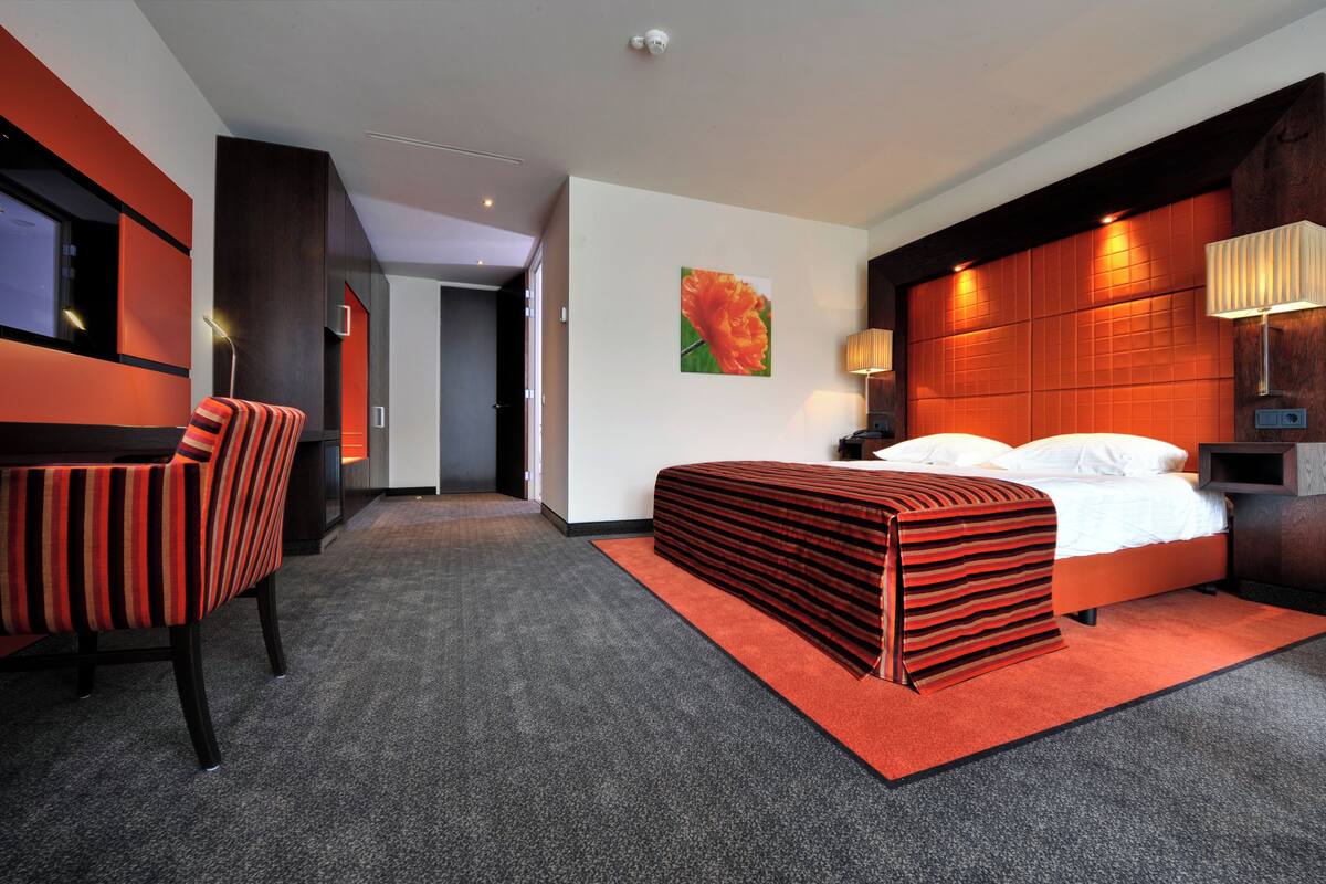A modern hotel room with a bed, a chair, a desk, and a painting on the wall.