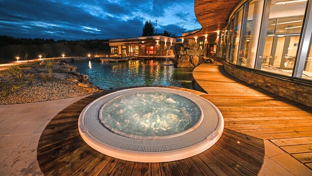 Un bain à remous avec une terrasse en bois, une piscine et un bâtiment doté de grandes fenêtres et d’une façade en pierre.